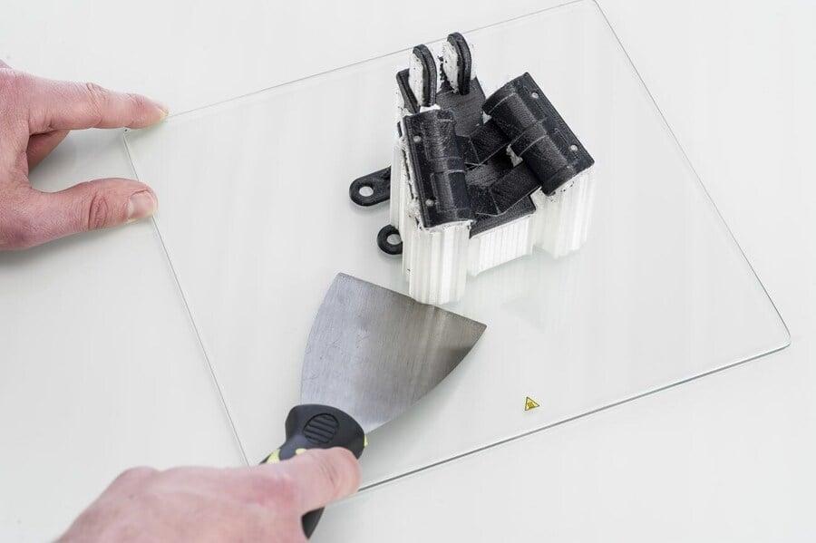 A spatula removes a 3D printed object from a glass print bed.