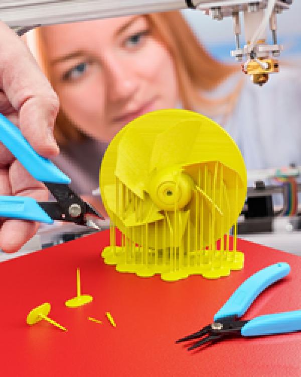 A woman in the background uses wire cutters to remove a 3D printed fan from the build platform.
