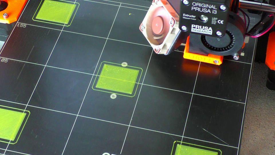 A 3D printer prints a green square on a black platform.