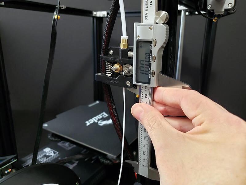 A hand is holding a digital caliper up to a Bowden tube connected to a 3D printer.