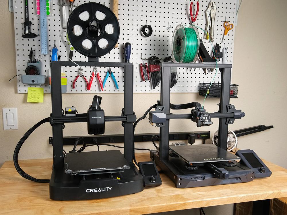 Two Creality 3D printers sit on a table in front of a pegboard wall with tools hanging on it.