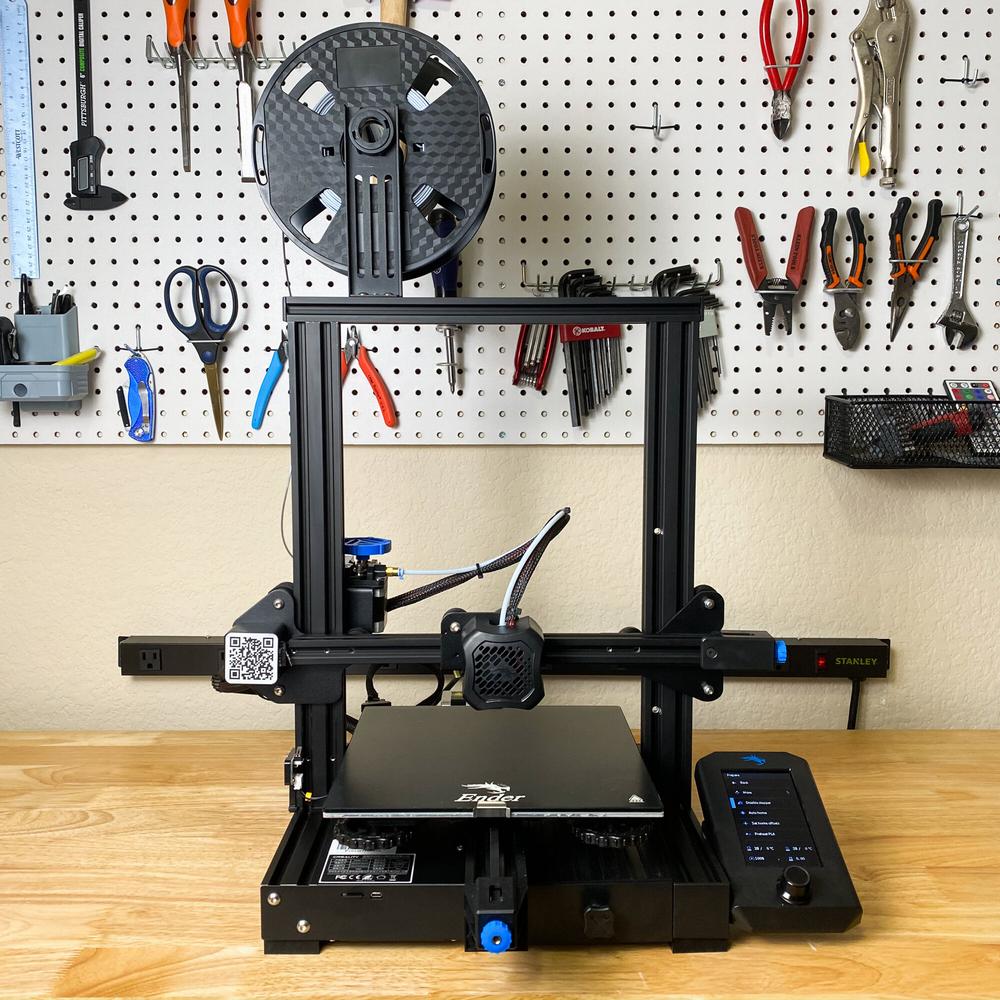 Black and blue Creality Ender 3 V2 3D printer on a wooden table in front of a pegboard wall with tools hanging on it.