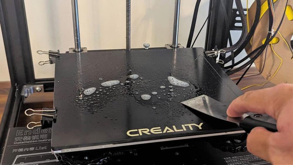 A person is cleaning the print bed of a Creality Ender 3 3D printer with a spatula.
