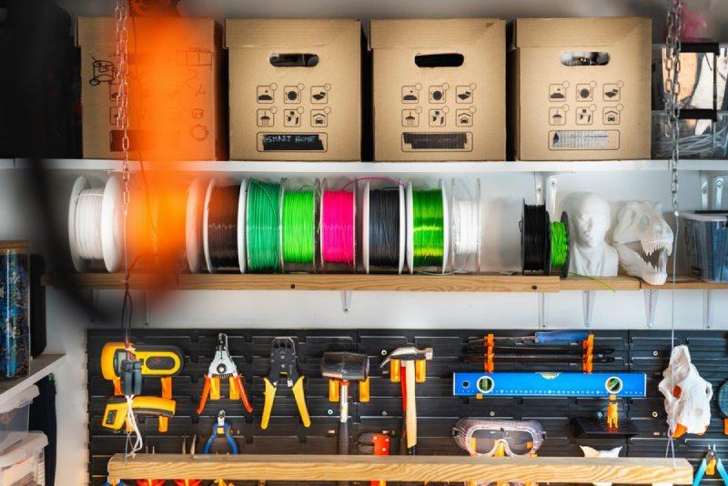 The image shows a garage shelf with various items on it, including spools of filament, a 3D printed dinosaur skull, and an assortment of tools.