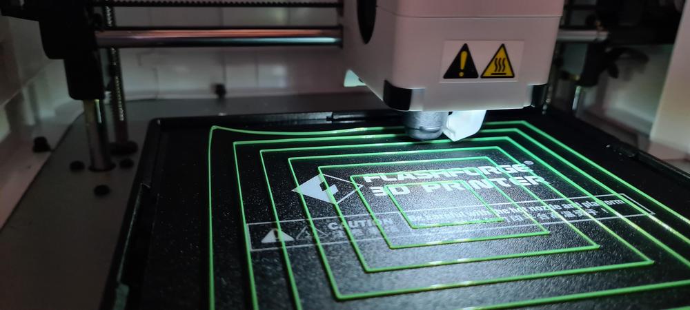 A 3D printer prints a green object on a glass bed.