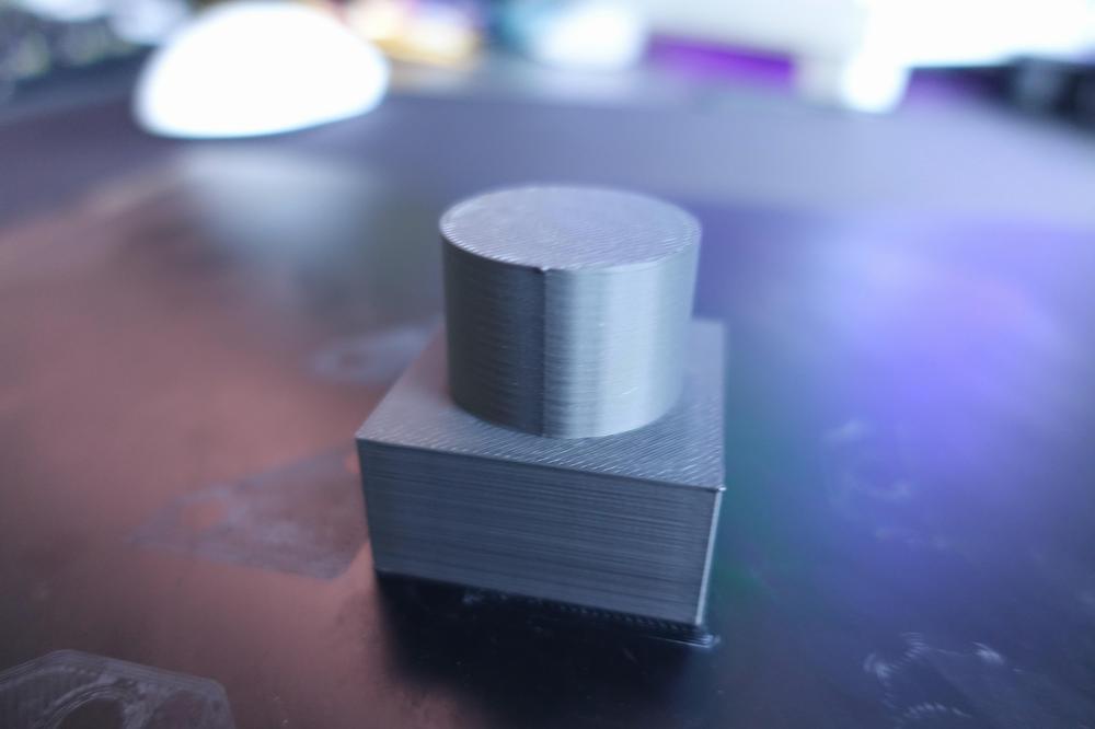 A black 3D printed cylinder sits on top of a black 3D printed square.