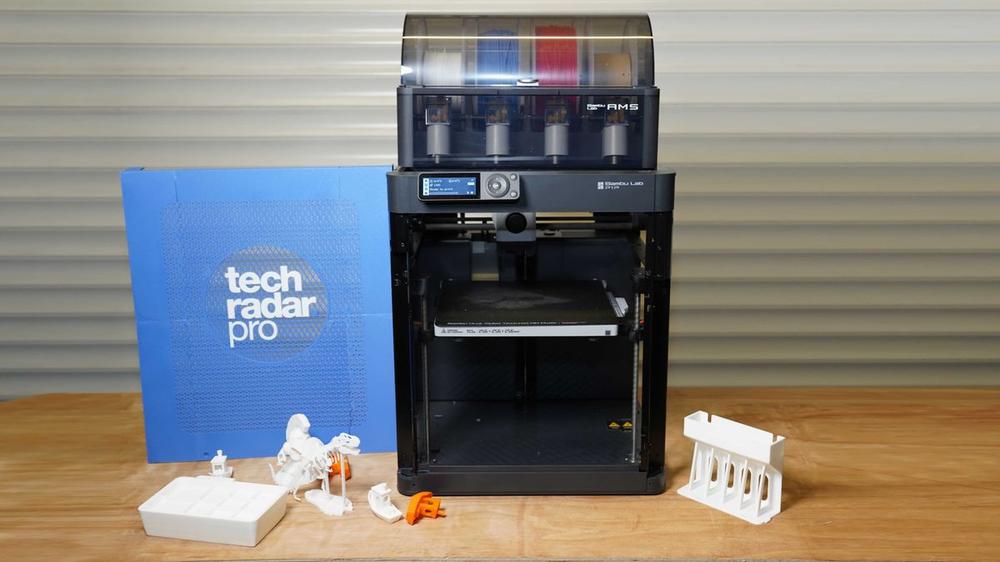 The image shows a black and grey 3D printer with a blue light on the front and a touchscreen, with a partially printed white dinosaur model on the print bed and several other white and orange printed models next to it.