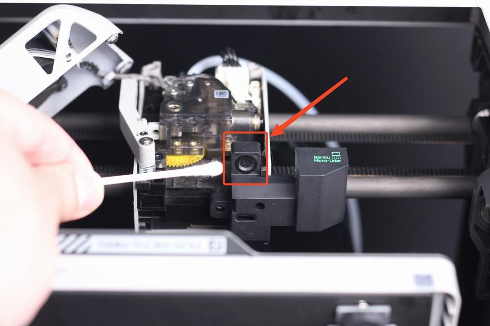 The image shows a person cleaning the nozzle of a 3D printer with a cotton swab.