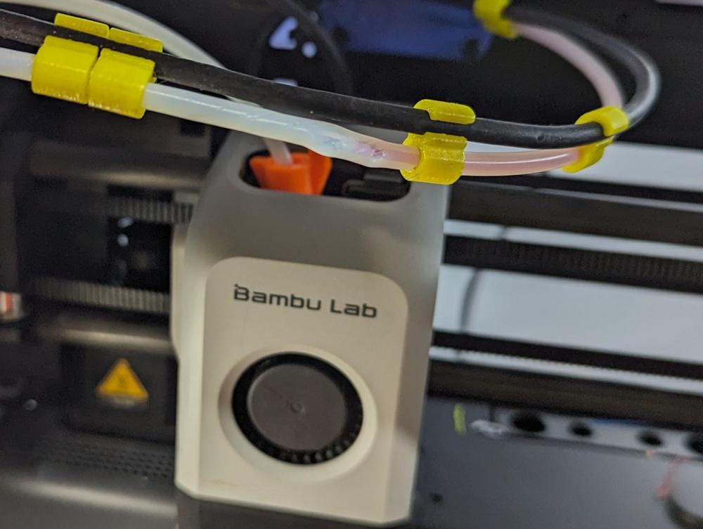 Close-up of a white and gray 3D printer with yellow cable ties holding the filament tube in place.