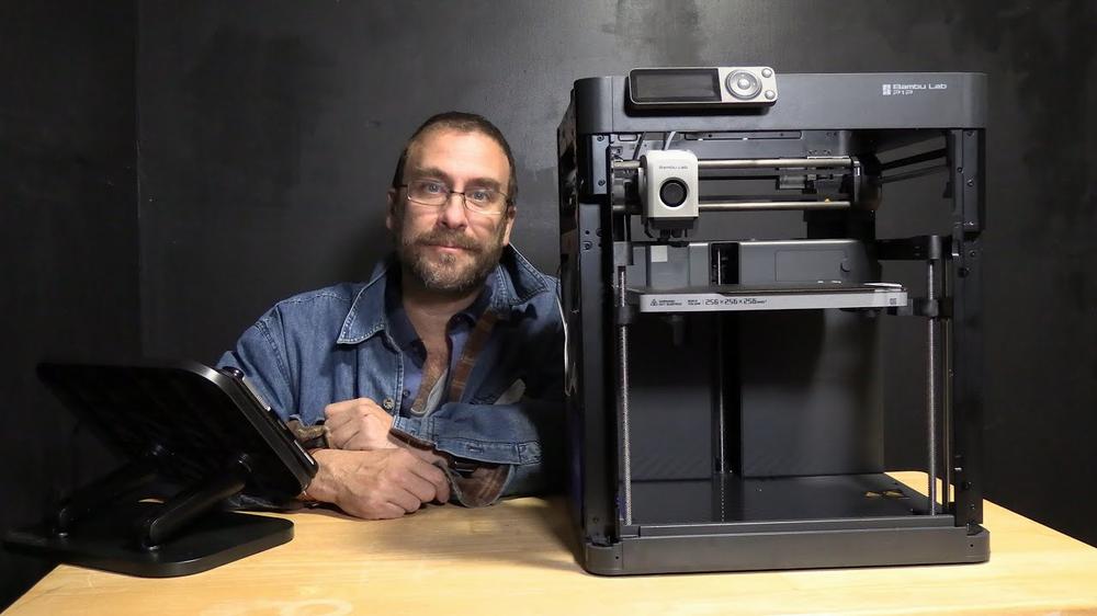 A man with glasses and a beard is standing next to a 3D printer.