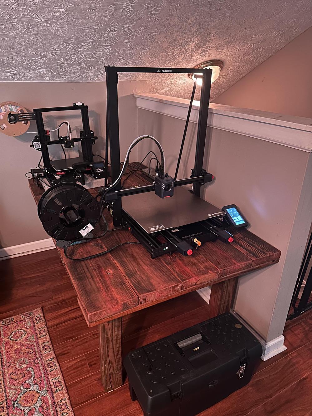 Two 3D printers sit on a wooden table in a room with a tan carpet and white walls.