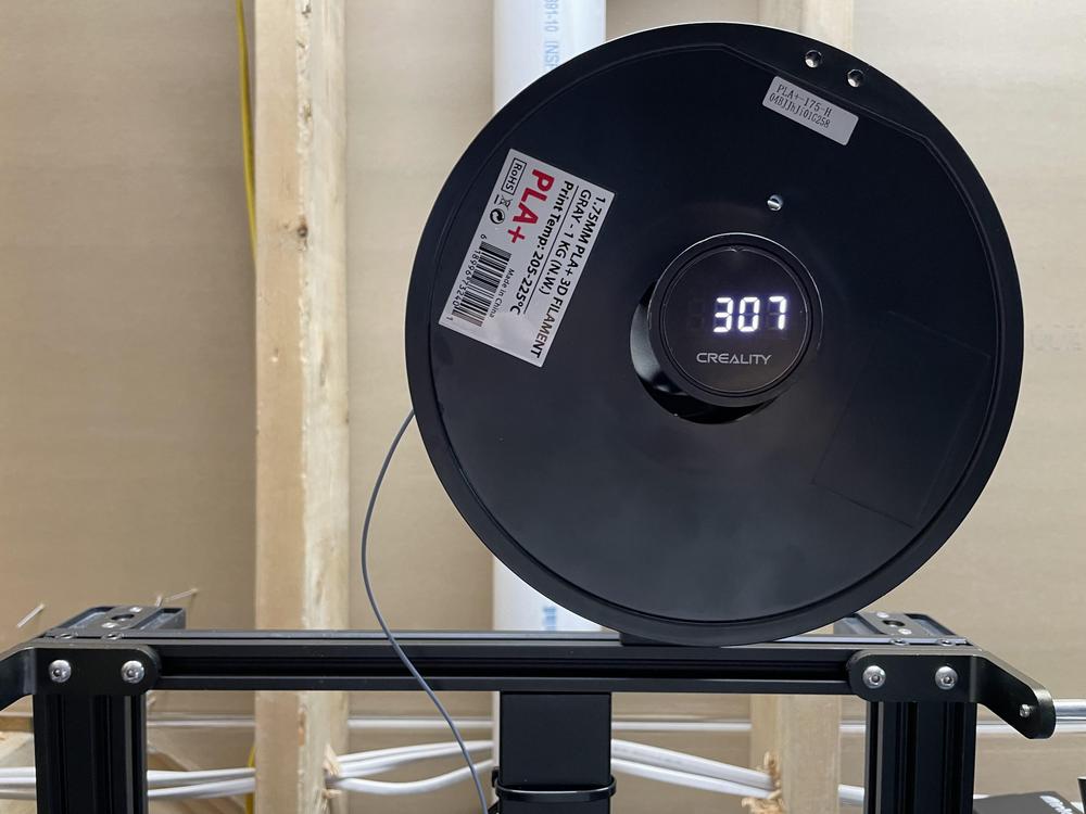 A black spool of PLA+ filament sits on a 3D printer with a label showing the filament type, size, and temperature.