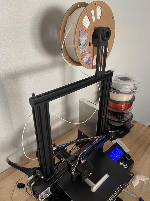 A white spool of 3D printer filament sits on a holder next to a black 3D printer.