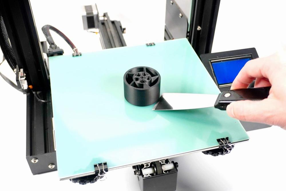 Black 3D printed object on a blue magnetic print bed, being removed with a spatula.