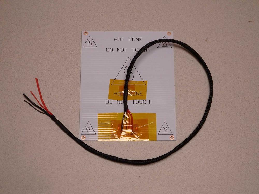 A black silicone heater pad with a thermistor wire attached to it, with a warning label saying Hot Zone, Do Not Touch!