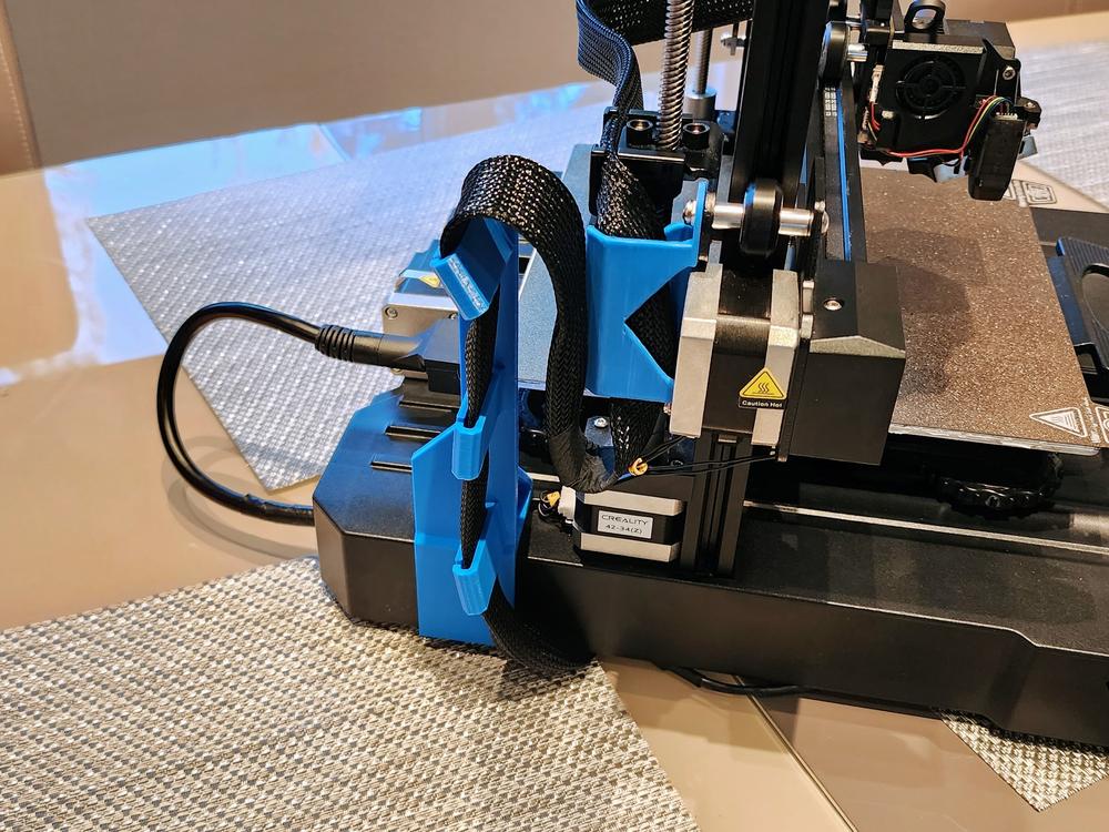 A blue 3D printed part is attached to the side of a Creality Ender 3 V2 3D printer, holding the power cable in place.