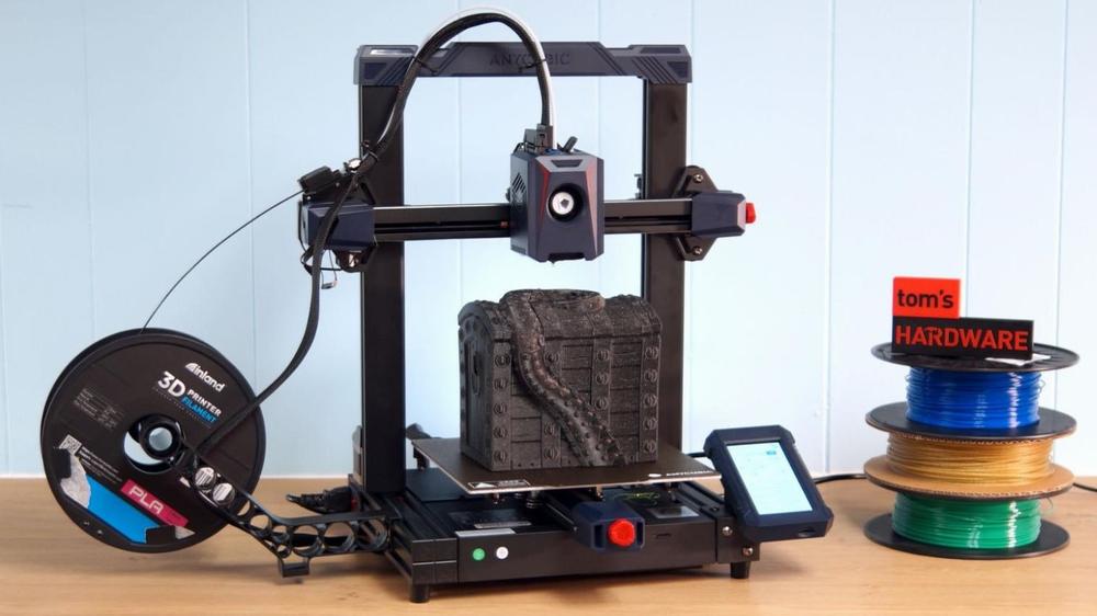 A black and red 3D printer is printing a grey object while spools of filament sit next to it.