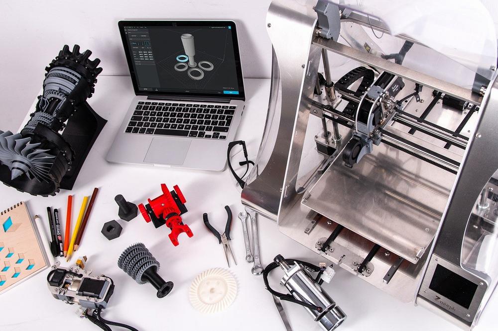 A 3D printer sits on a table next to a laptop and various tools and parts.