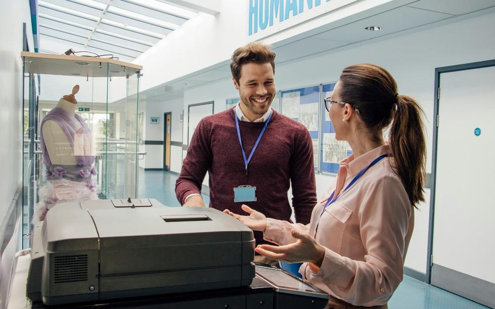 A man and a woman are standing next to a printer, talking and smiling.